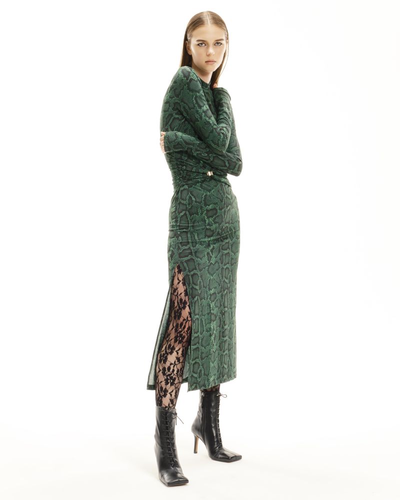 A woman poses in a full-length shot, wearing a green snake-print dress with a slit, black lace tights, and black lace-up heeled boots on a white backdrop.
