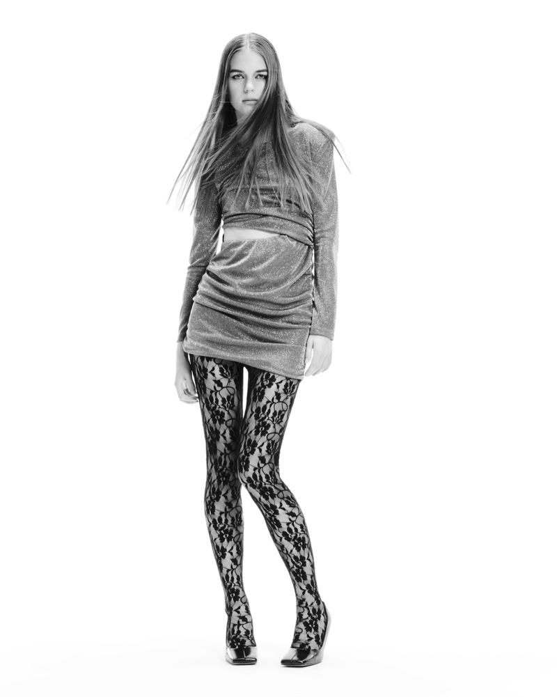 Model Tilda Jönsson wearing a grey long-sleeve mini dress and black floral lace tights against a white studio background.