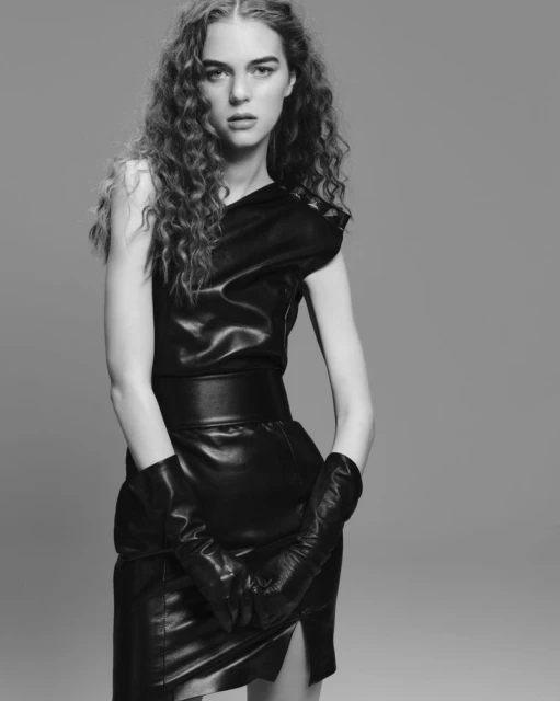 Monochrome studio shot of a stylish adult female model in a black leather dress and gloves, against a neutral gray backdrop.