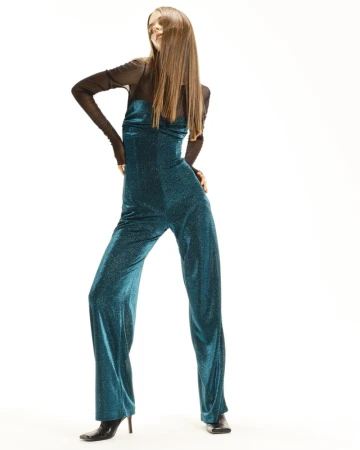 Model Tilda Jönsson posing in a shimmering teal jumpsuit layered over a sheer black top against a white studio background.