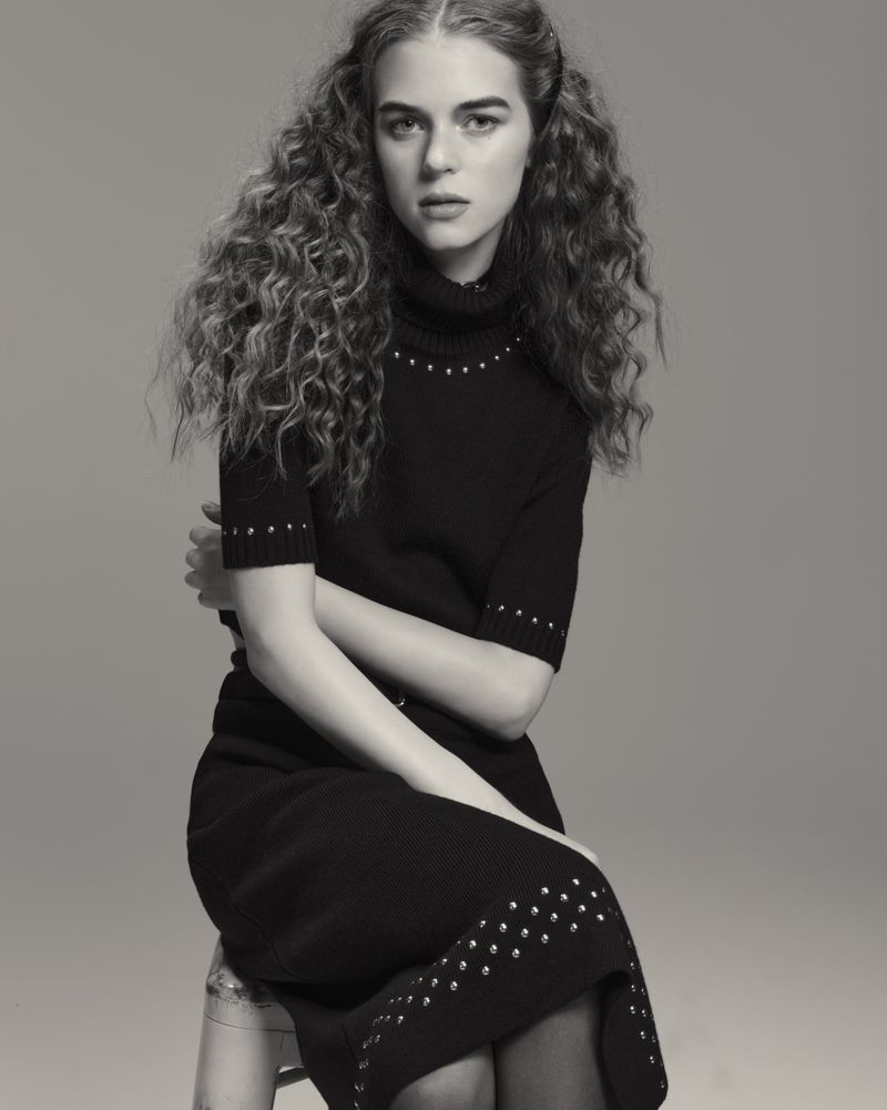 Monochromatic portrait of a curly-haired model in a black dress sitting on a clear stool in a photo studio with a neutral background.