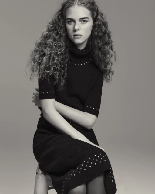 Monochromatic portrait of a curly-haired model in a black dress sitting on a clear stool in a photo studio with a neutral background.