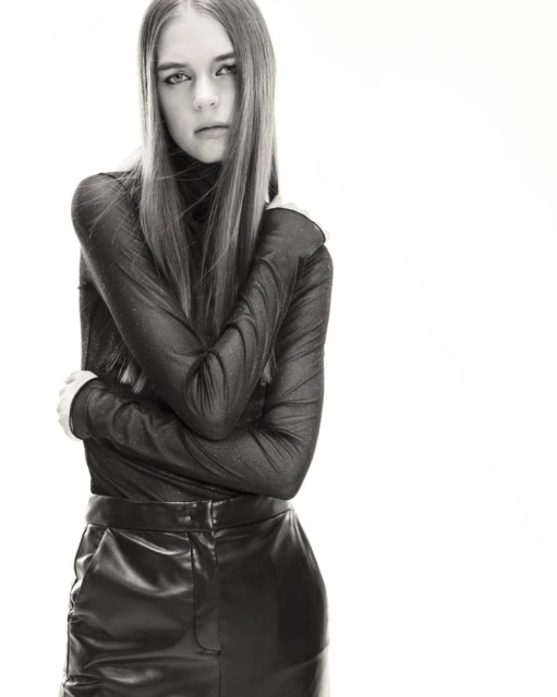 Portrait of a young adult female model in a long-sleeved black top and black faux-leather skirt, posing indoors against a white background.