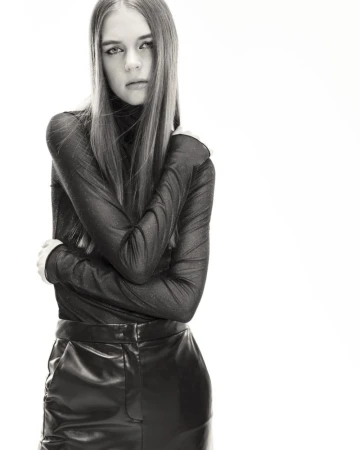 Portrait of a young adult female model in a long-sleeved black top and black faux-leather skirt, posing indoors against a white background.
