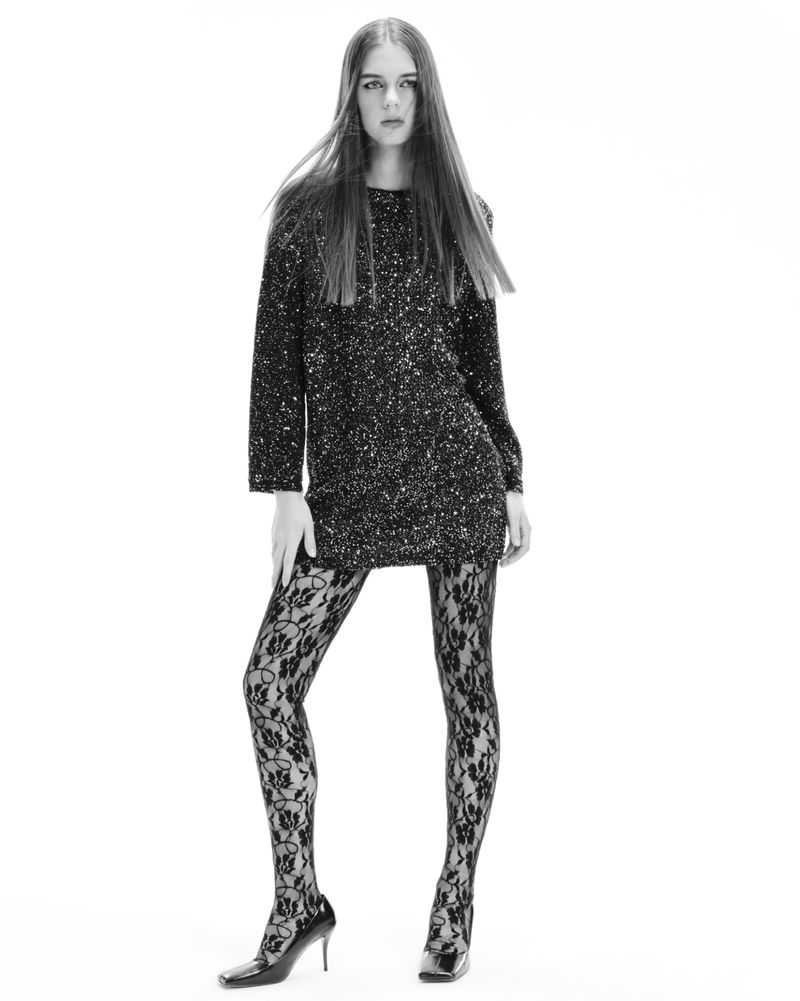 Full-length studio shot of a young adult woman with long brown hair, wearing a sparkly dress, floral tights, and heels on a white background.