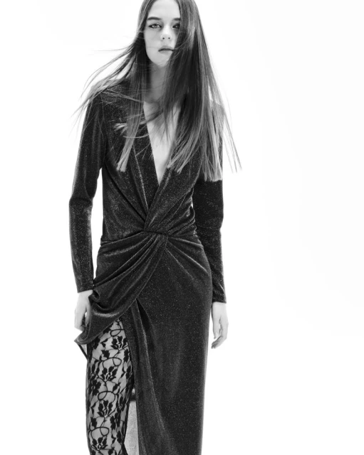 Black and white fashion shot of an adult female model in a long-sleeve dress with a thigh-high slit, posing in a photo studio.