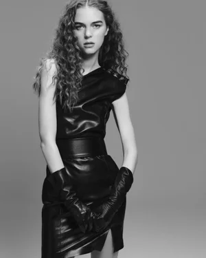 Model Tilda Jönsson in a black sleeveless leather dress and matching long leather gloves against a grey studio backdrop.