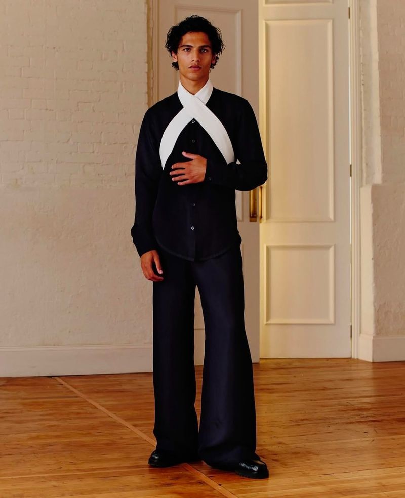 Man poses in black shirt and wide-leg pants, accessorized with a crossed white sash, standing against neutral wall and door backdrop.