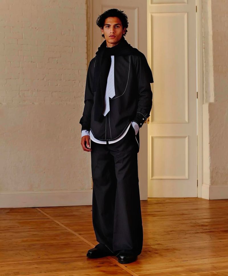 A person in a black and white pinstripe outfit with a scarf, posing indoors against a neutral background and wooden floor, medium shot.