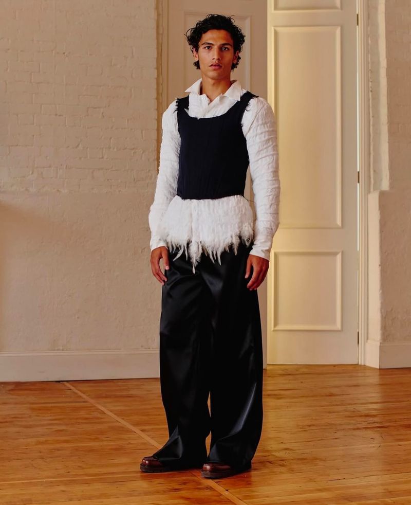 Standing fashion model in a white long-sleeved shirt, black vest, and shiny black wide-leg pants in a minimalist interior setting.