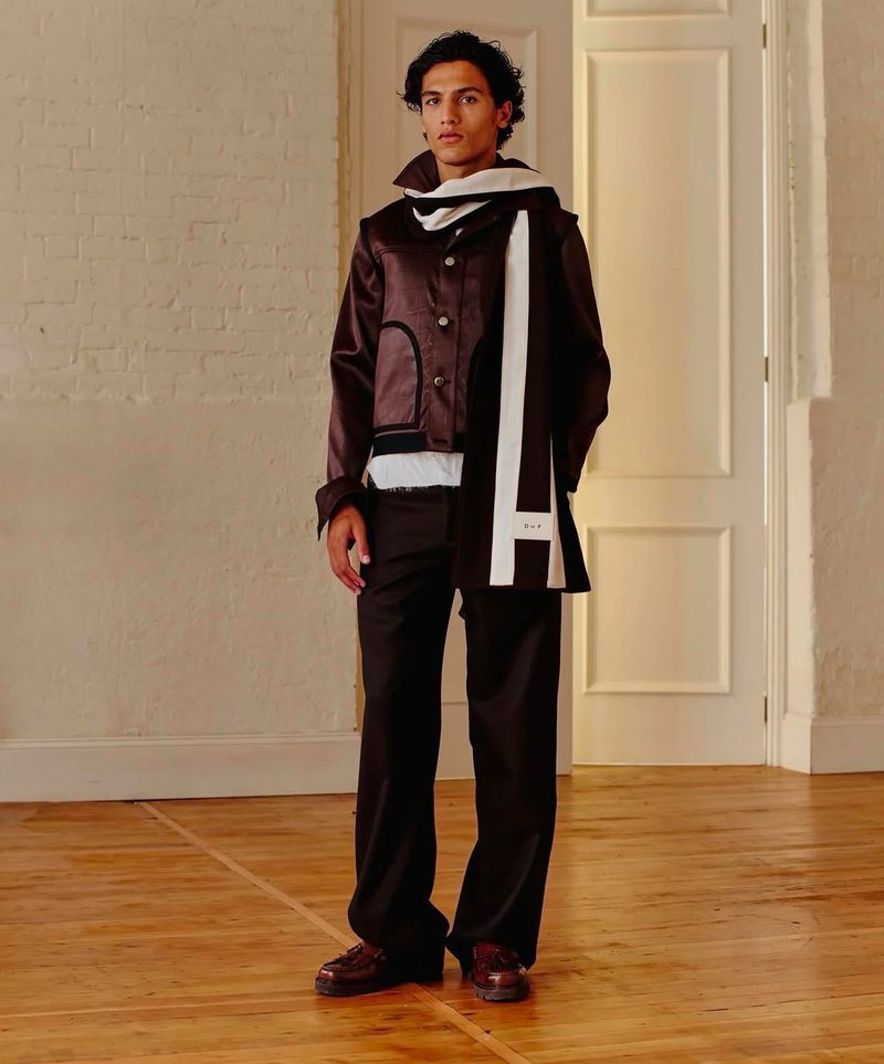 Fashionable young man in brown jacket and pants, accessorized with a white scarf in an indoor setting with neutral background.