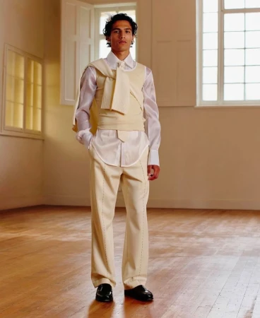 Male fashion model in off-white suit with draped sweater stands in naturally lit room, posing for a fashion editorial or advertisement.