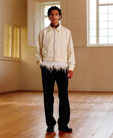 Male model stands in cream jacket and black pants in a minimalist space with wood floors and natural light from adjacent windows.