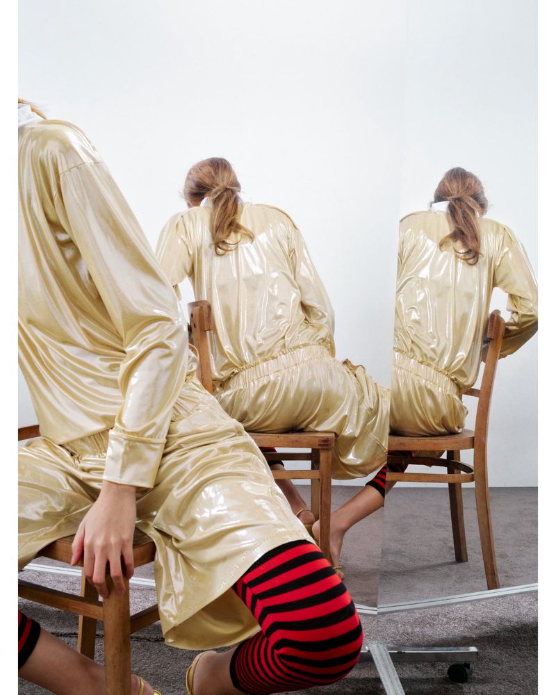 Three back views of a person sitting on a wooden chair wearing a shiny gold long-sleeve top and red and black leggings in a white studio.