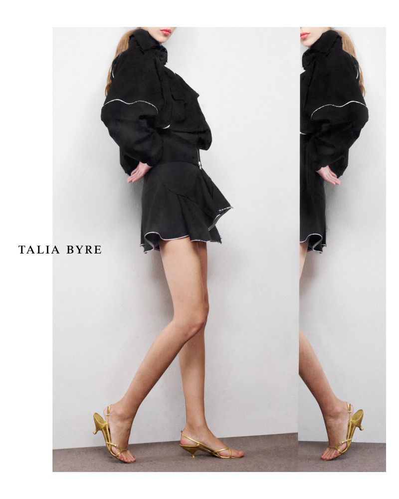 Fashion shot of a woman in a black jacket and skirt, paired with gold sandals against a plain background, promoting Talia Byre.