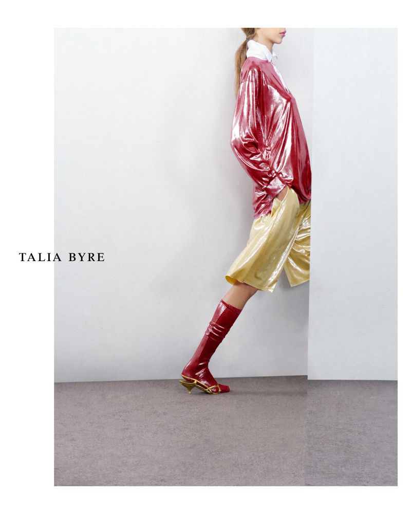 Fashion editorial shot: model poses in a pink metallic top, gold shorts, and red patent leather boots against a white backdrop.