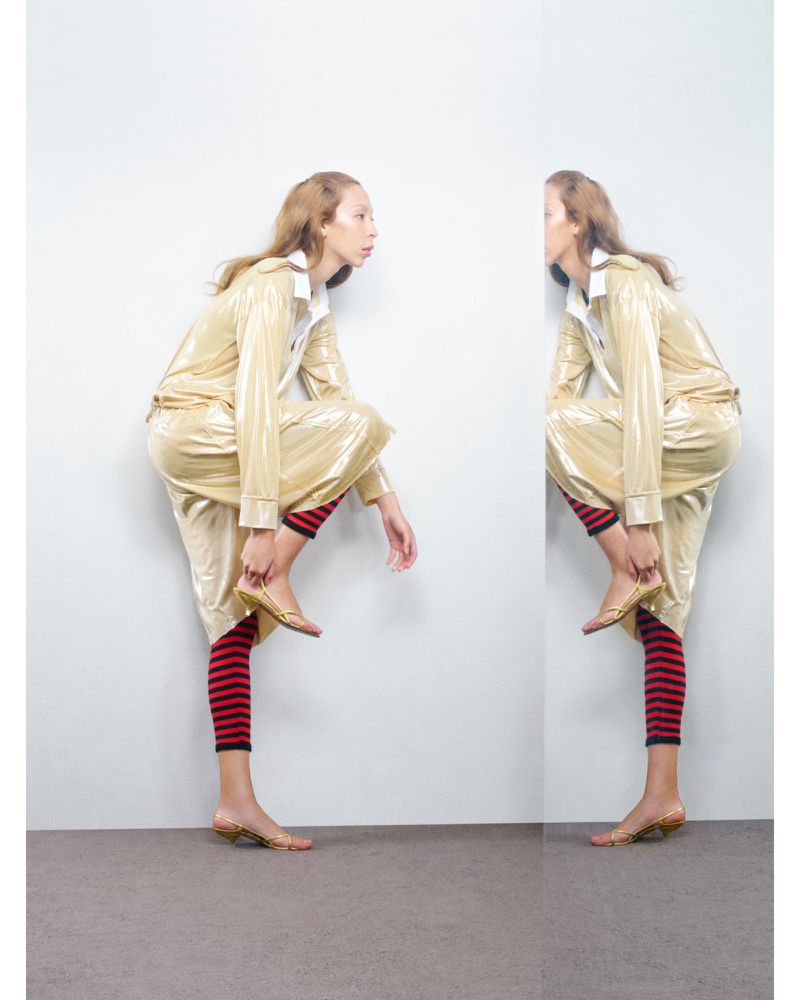 Fashion shot of a woman in a gold pyjama-style suit, striped leggings, and heels, posing beside a wall that creates a mirror effect.