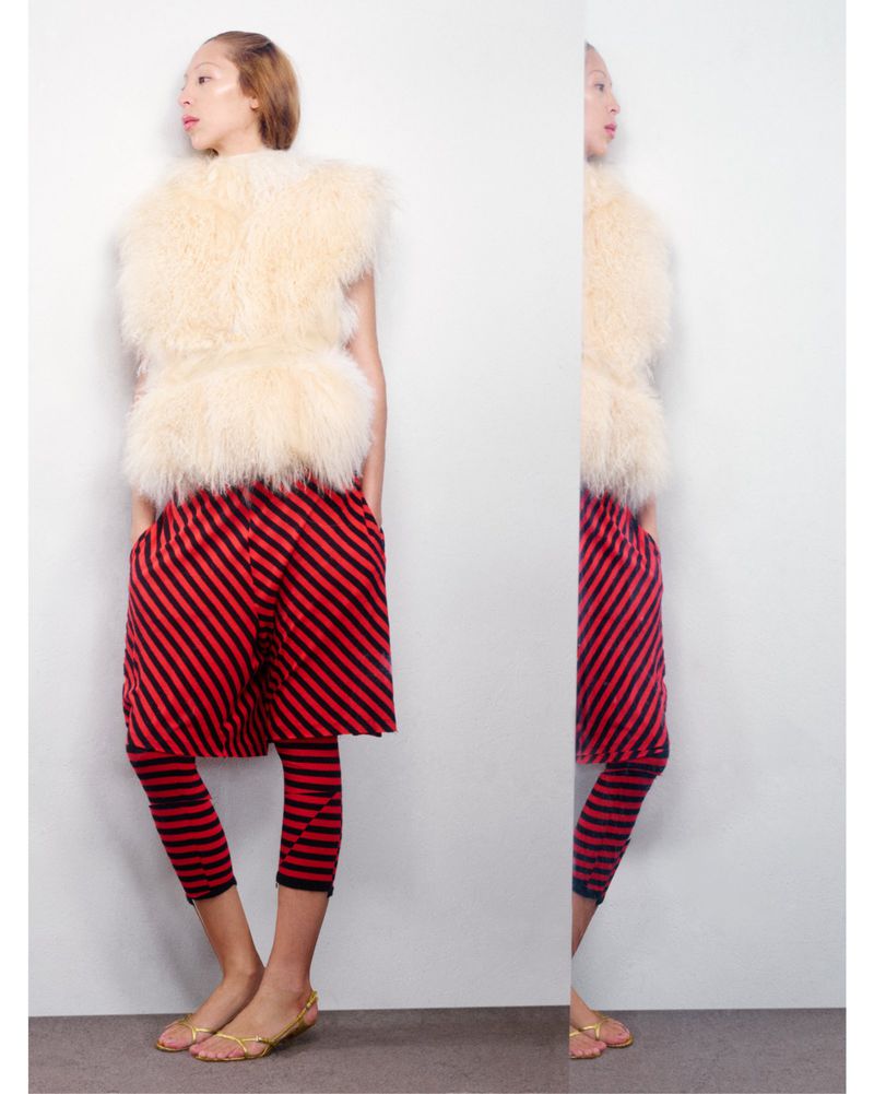 Fashion shot: woman poses in fur vest and red-striped pants against a white wall. Studio lighting. Fashion editorial style.