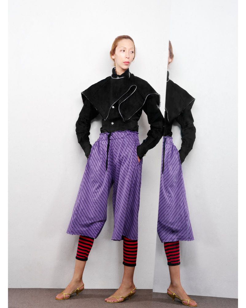 A slim woman with fair skin poses in a studio wearing culottes and a black jacket. There is a reflection of her in the background.