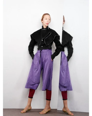 A slim woman with fair skin poses in a studio wearing culottes and a black jacket. There is a reflection of her in the background.