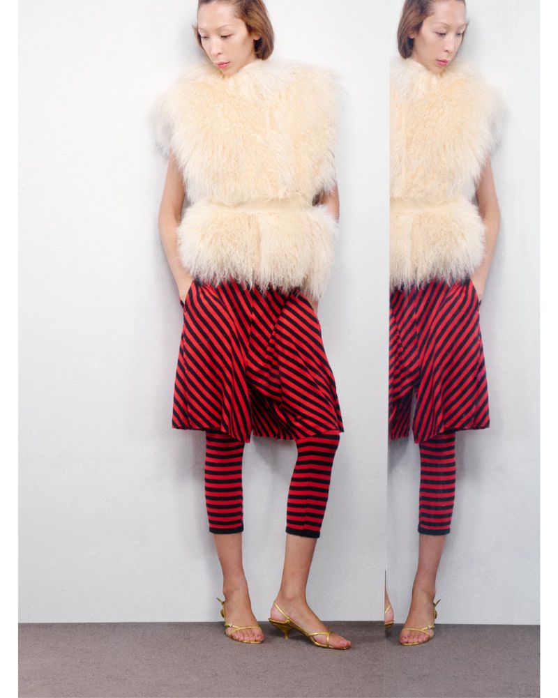 Woman in a fur vest, skirt, cropped leggings with gold sandals poses in front of a light gray wall in a studio fashion shot.