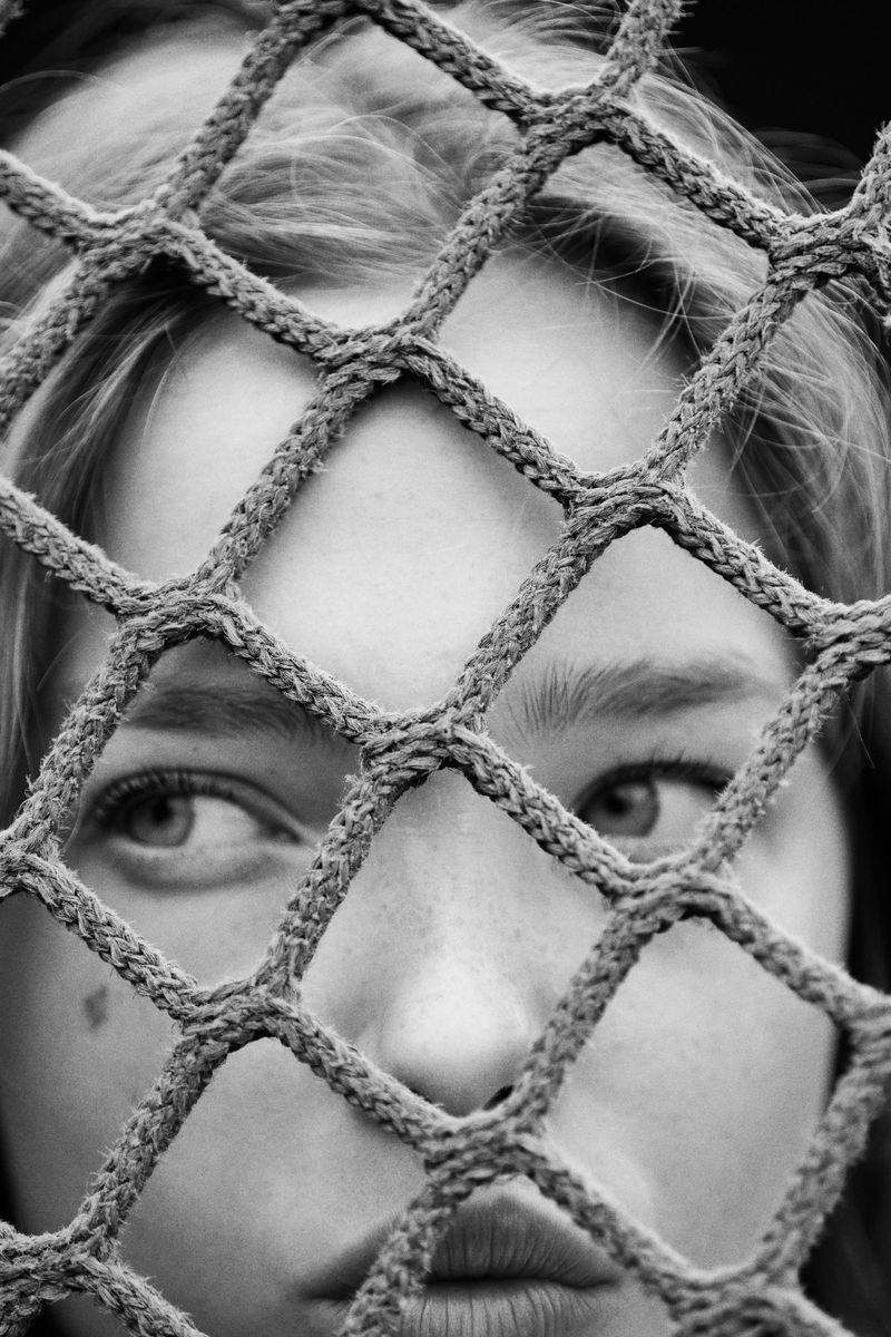 Close-up black and white portrait of a fair-skinned adult female partially obscured by a diamond-patterned netting in a neutral setting.