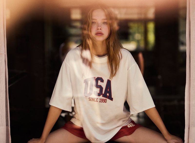 Blonde woman wearing a white USA t-shirt and red shorts poses indoors with soft lighting and blurred background elements.