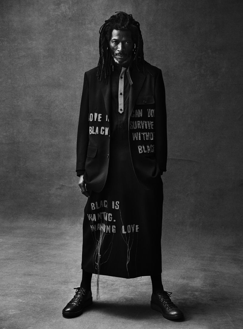 Black and white full shot of a man with dreadlocks in black jacket and dress with white text, standing against a grey backdrop.