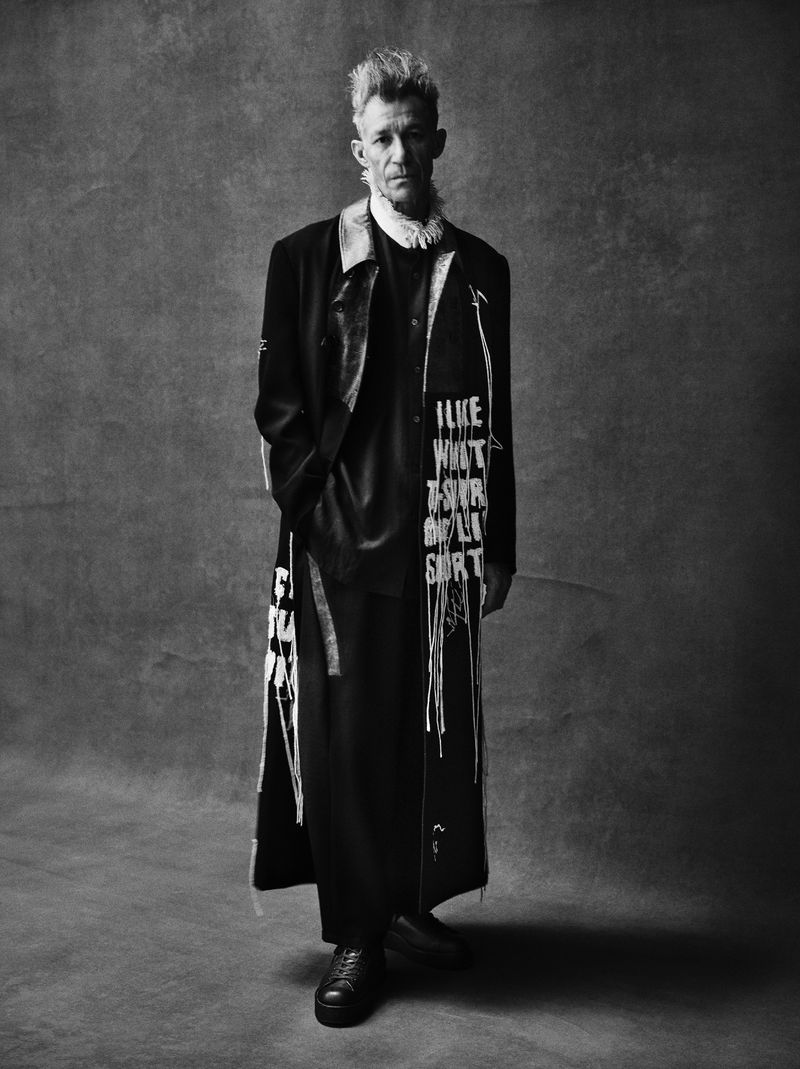 Full-length black and white studio portrait of a man with spiky blond hair wearing an overcoat with text print and black dress shoes.