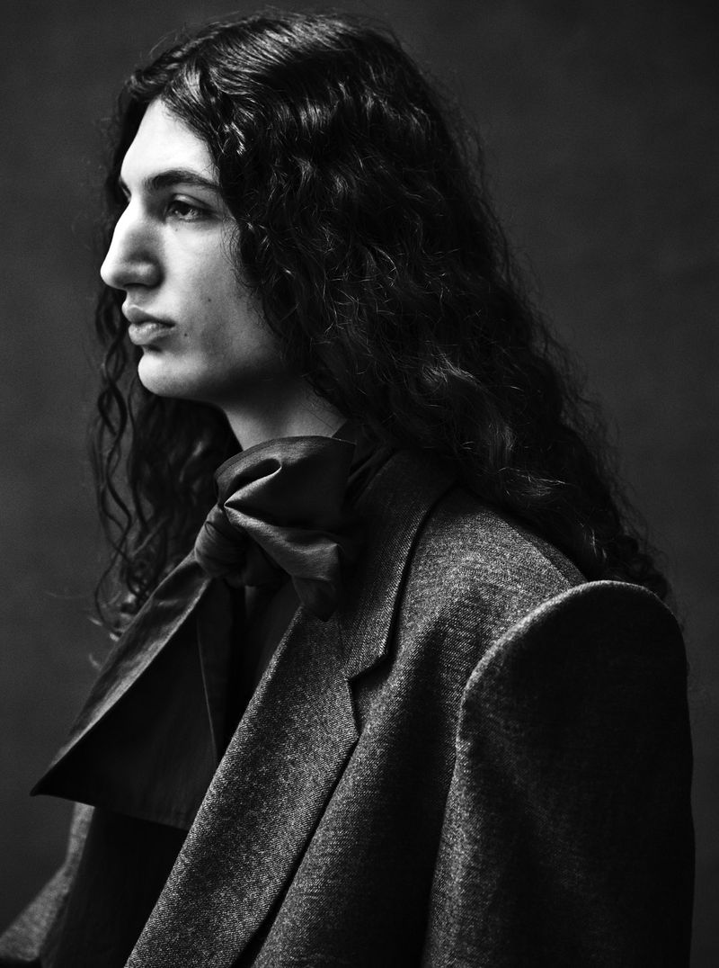 Monochrome portrait of a male with long dark curly hair in profile, wearing a dark bow and jacket against a dark background.