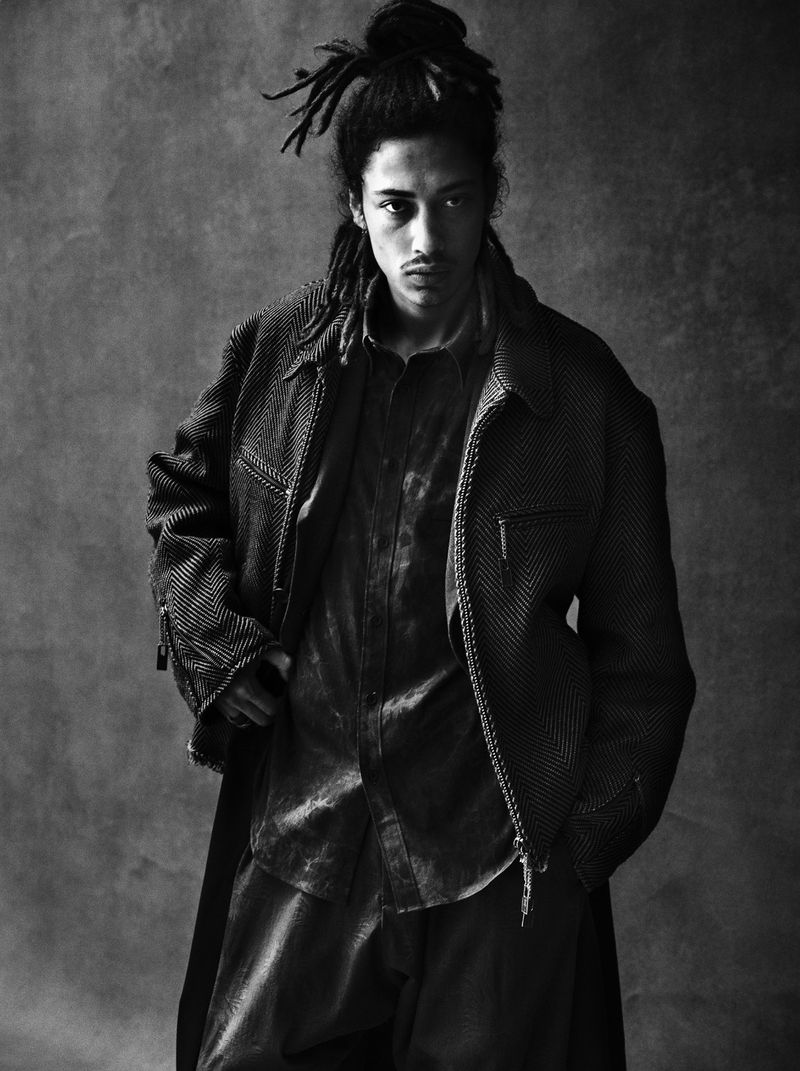 Monochrome portrait of a male model with dreadlocks wearing an open jacket and collared shirt against a grey mottled backdrop.