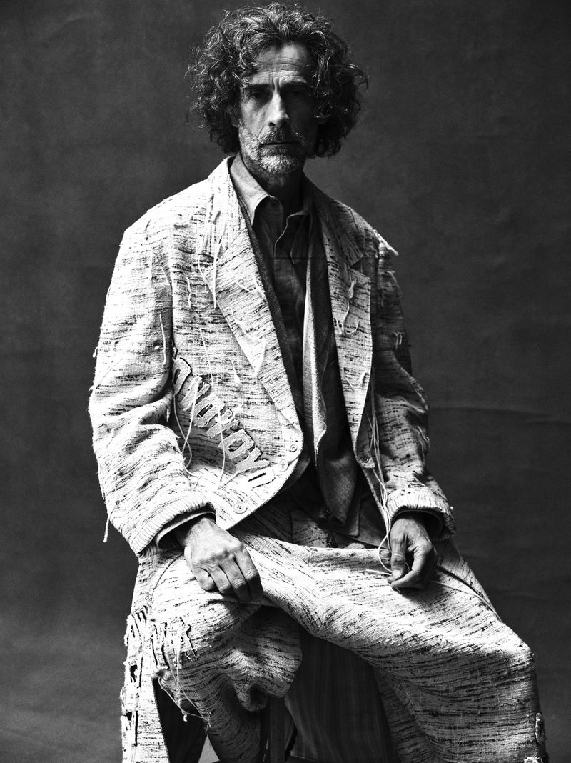 Black and white portrait of a mature adult man with curly hair, wearing a textured suit while seated against a dark backdrop.