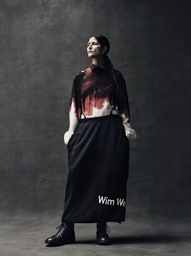 A woman in a dark skirt and reddish top poses against a gray background, looking up and away from the camera in a fashion shot.