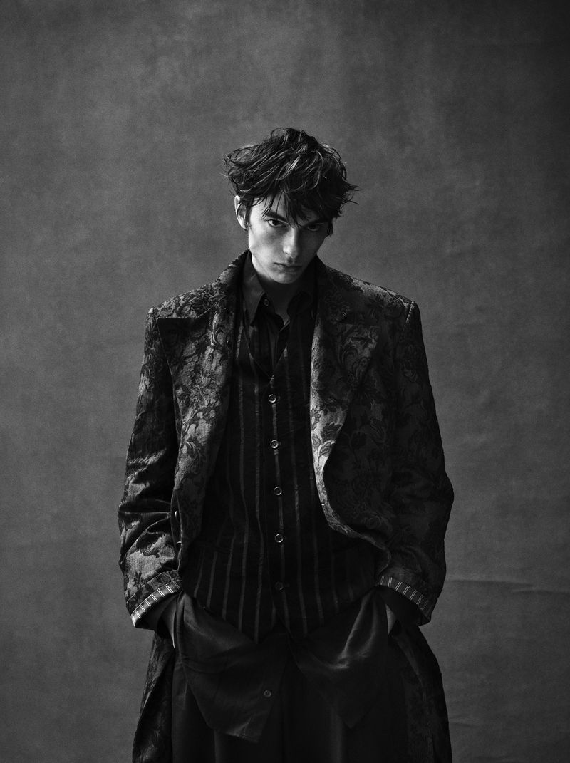 Black and white portrait of a person in a patterned jacket and striped shirt, posing indoors against a textured backdrop, hands in pockets.