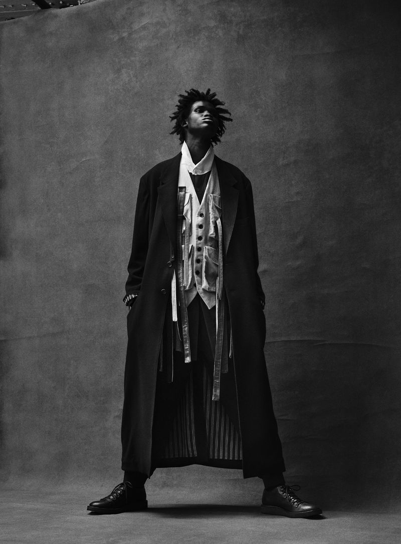 Monochrome full shot of an adult male with dreadlocks wearing a long black coat, vest, and dress shoes, standing against a grey background.