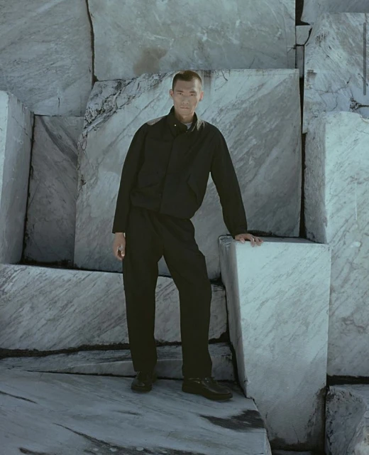 Full shot of an adult man wearing a black jacket and pants, standing next to a large block of marble with visible veining and texture.