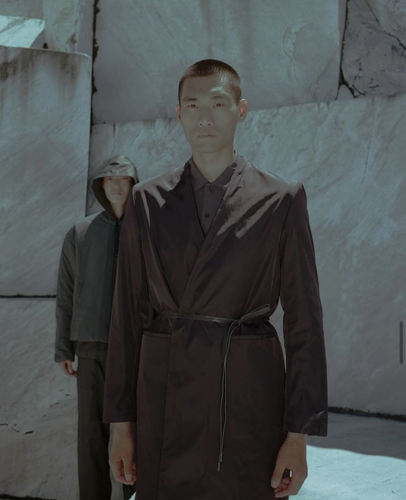 Two males in matching minimalist clothing, one in an open overcoat tied with a cord, pose against a rough-hewn gray marble wall.