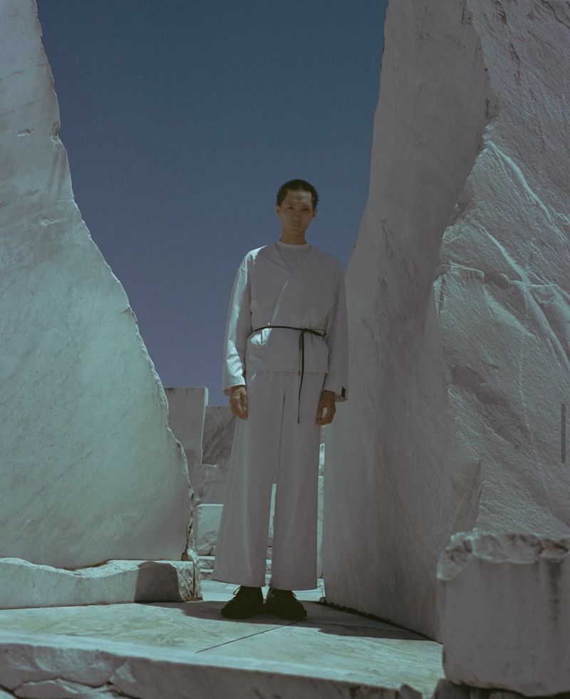 Adult male in light-colored fashion, posing between light marble-like slabs, wearing a robe-like garment with a thin, dark sash.