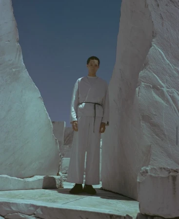 Adult male in light-colored fashion, posing between light marble-like slabs, wearing a robe-like garment with a thin, dark sash.