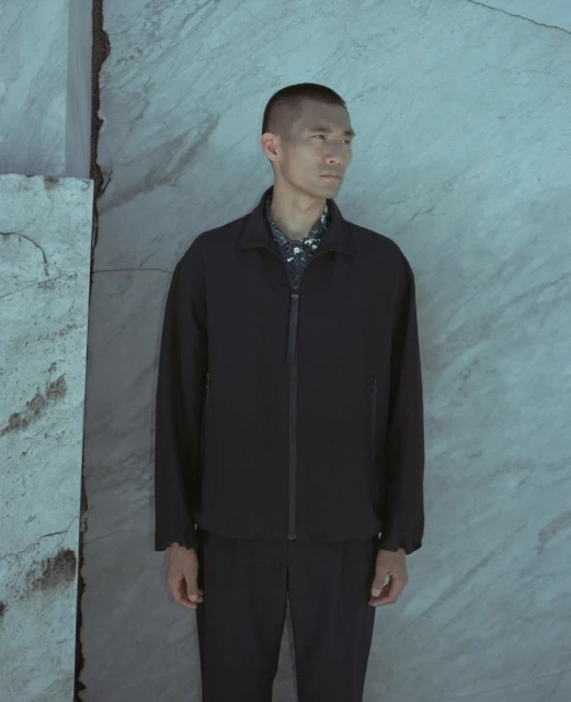 Asian man in dark jacket and pants posing against a mottled stone wall, looking to the side. Dark navy-blue clothing against mottled stone backdrop.