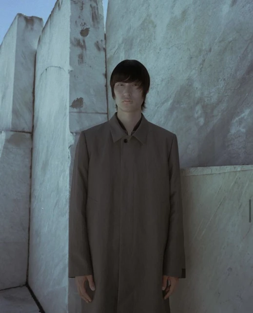 Portrait of a slender adult with a neutral expression, wearing a brown coat, posing in front of geometric textured marble walls.