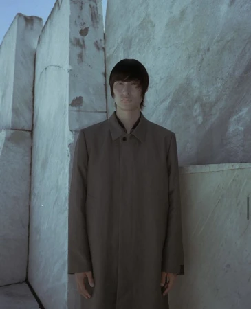 Portrait of a slender adult with a neutral expression, wearing a brown coat, posing in front of geometric textured marble walls.