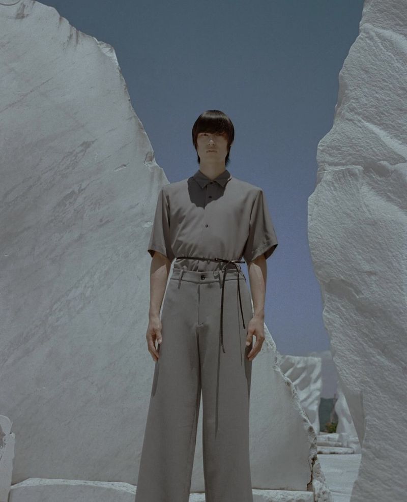 A person in muted tones stands amidst towering white rock formations against a pale blue sky in a fashion editorial setting.
