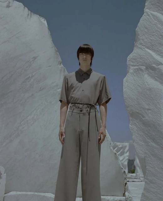 A person in muted tones stands amidst towering white rock formations against a pale blue sky in a fashion editorial setting.