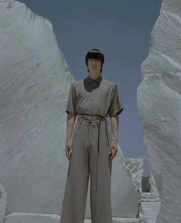 A person in muted tones stands amidst towering white rock formations against a pale blue sky in a fashion editorial setting.