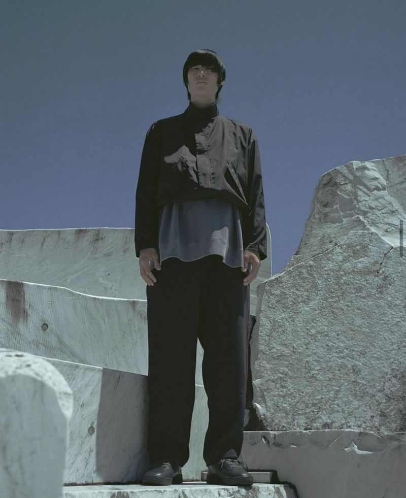 A person standing on stone steps outdoors with a blue sky background, wearing a black jacket and pants in a full shot.