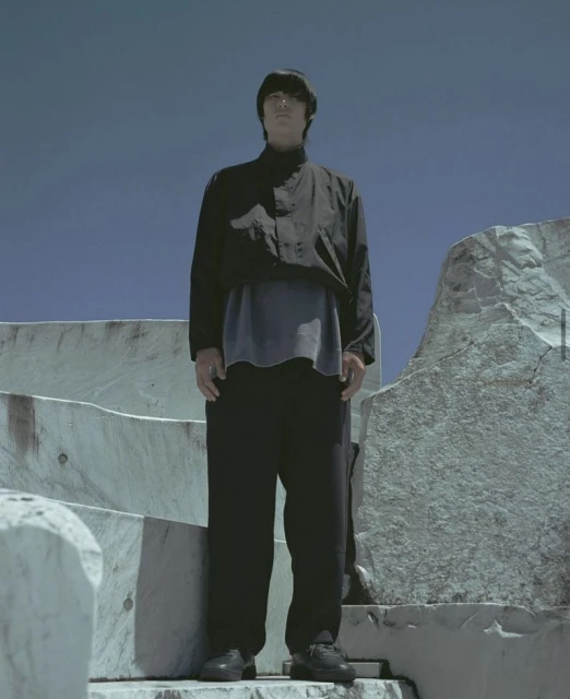 A person standing on stone steps outdoors with a blue sky background, wearing a black jacket and pants in a full shot.
