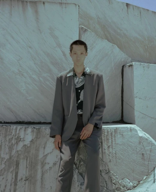 A fashionable adult in a grey suit stands against a bright stone wall. The subject is well-lit with a neutral expression and short hair.