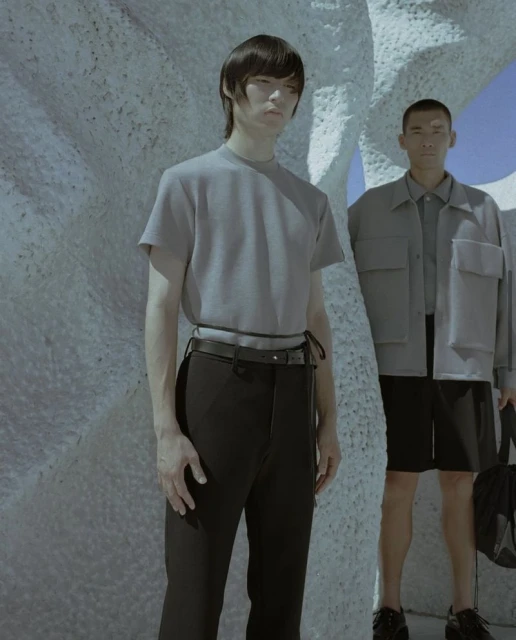 Two Asian male models pose outdoors in a studio fashion shoot featuring neutral color palettes and modern casual wear against a textured backdrop.