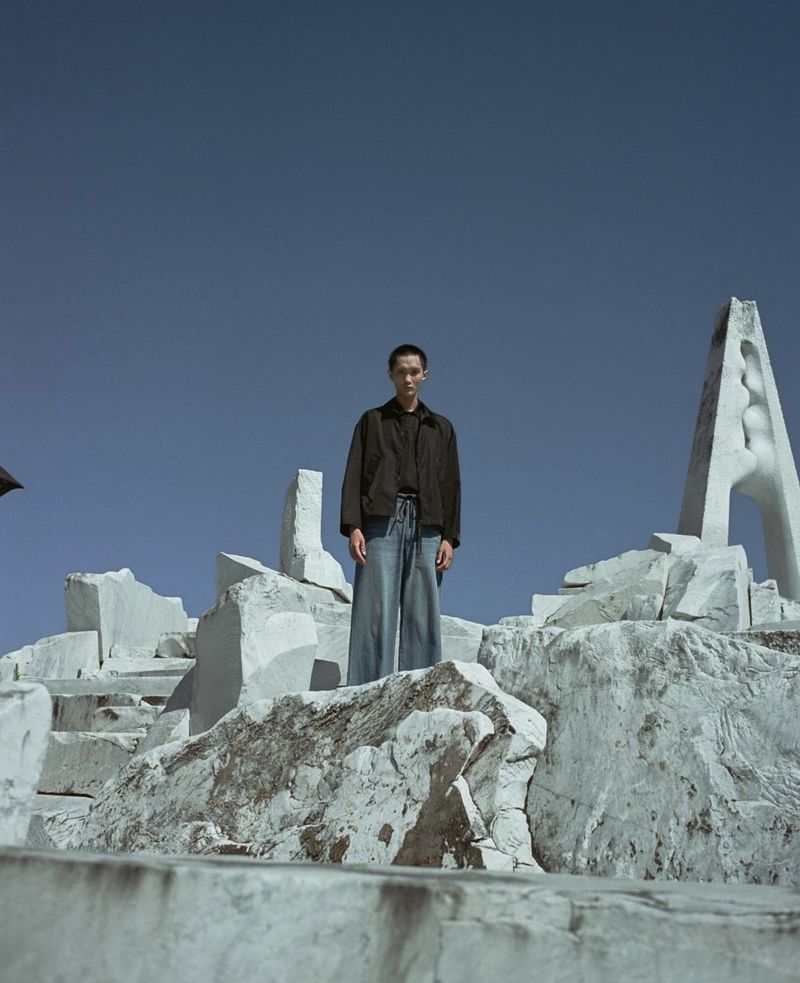 A person stands on a marble structure against a blue sky. The subject wears jeans and a dark jacket in an outdoor setting.